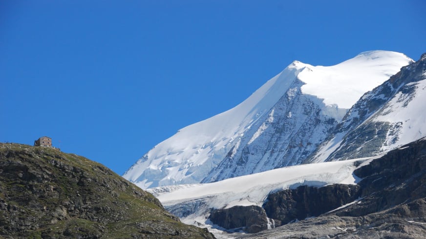 Die Turtmannhütte steht auf einem grasbewachsenen Hang vor verschneiten Gipfeln und einem Gletscher mit Spalten.