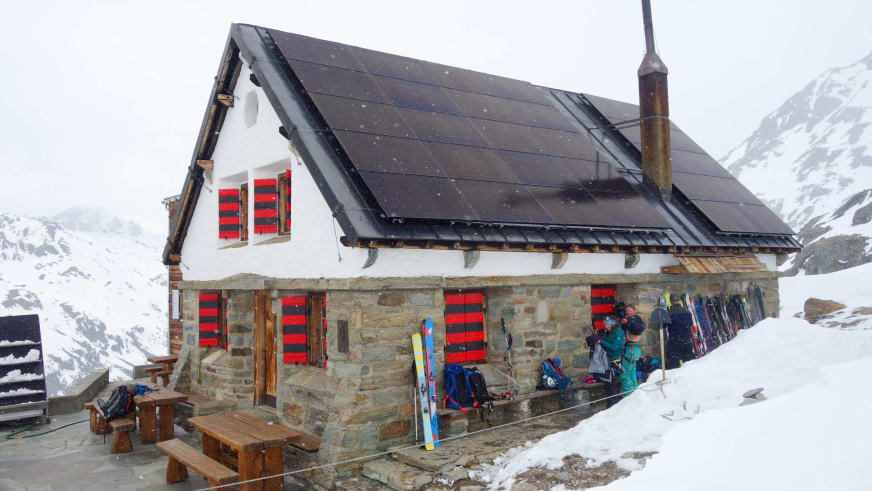 Skitourengeher machen Pause vor der Turtmannhütte, während Schneeflocken auf die verschneite Berglandschaft fallen.