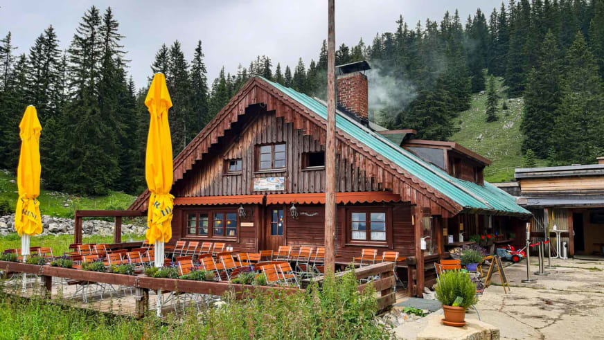 Außenansicht der Oberen Maxlraineralm mit überdachter Terrasse und rauchenden Schornstein im Almgebiet.