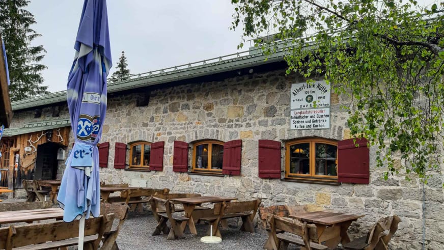 Sonniger Gastgarten der Albert-Link-Hütte mit Holzbänken und Sonnenschirm vor dem Natursteingebäude.