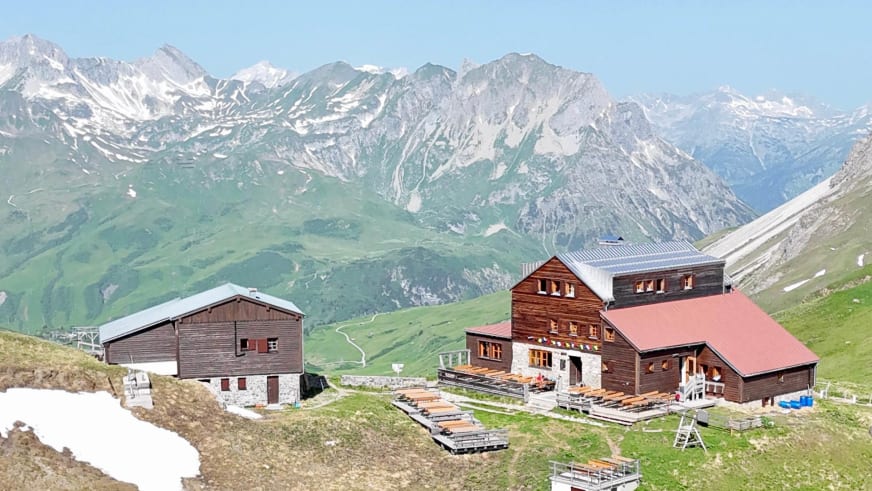 Panoramablick auf die Stuttgarter Hütte mit traditioneller Holzbauweise vor schneebedeckten Gipfeln der Lechtaler Alpen.
