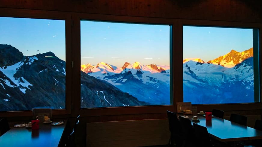 Gastraum der Hohsaashütte mit großen Fenstern und Panoramablick auf verschneite Berggipfel im Abendlicht.