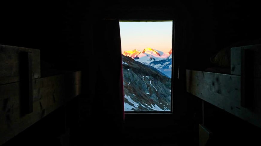 Blick durch ein Fenster der Hohsaashütte auf schneebedeckte Berggipfel im warmen Morgenlicht.