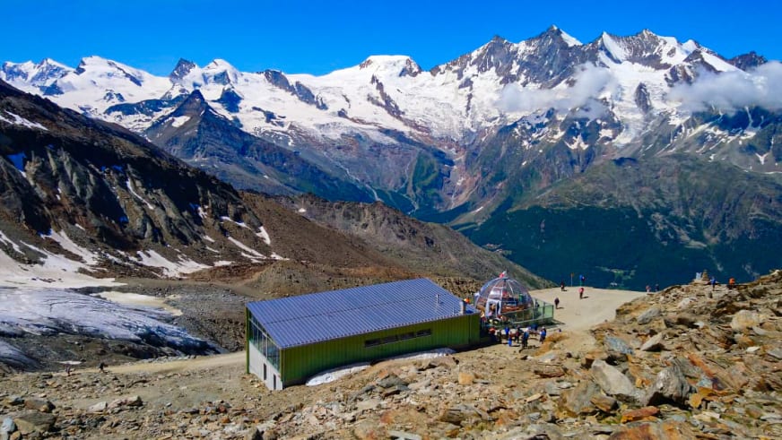 Luftaufnahme der Hohsaashütte mit grünem Gebäude und angrenzender Seilbahnstation auf steinigem Hochplateau.