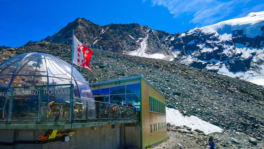 Die Hohsaashütte mit moderner Glaskuppel-Architektur steht auf steinigem Geröll vor schneebedeckten Berggipfeln.