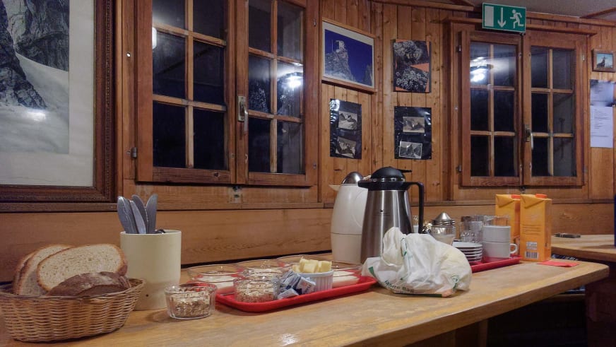 Frühstücksbuffet der Cabane de Bertol mit Thermoskannen und Brotkorb auf Holzbank.