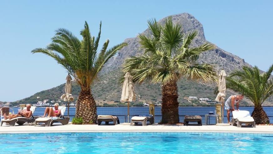 Poolbereich des Hotel Plaza Kalymnos mit Palmen, Liegestühlen und Blick auf die blaue Meeresbucht.