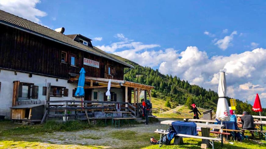 Wanderer versammeln sich vor der Galflunalm mit großer Sonnenterrasse und bunten Schirmen auf grüner Almwiese.