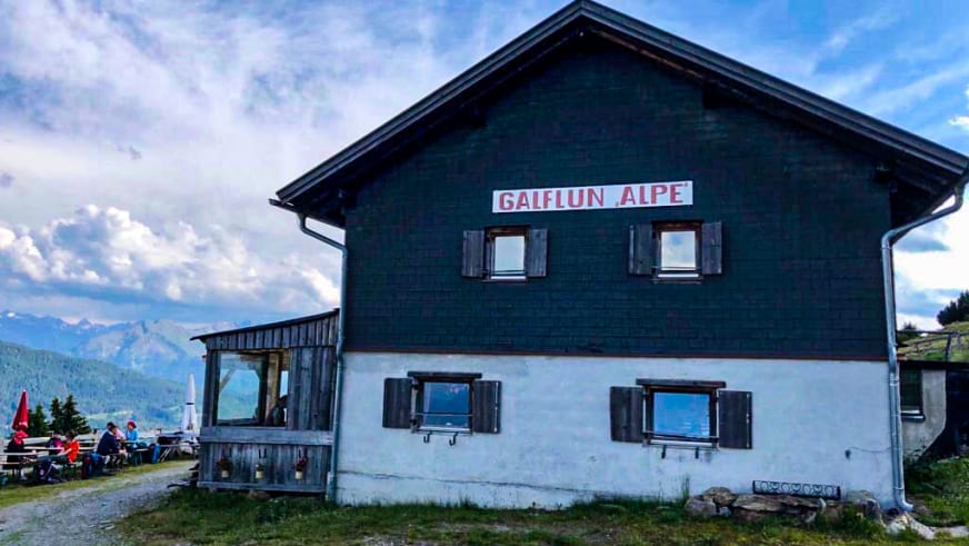 Die Galflunalm zeigt typische Alpenarchitektur mit Holzverkleidung und weißem Erdgeschoss sowie Terrasse mit Gästen.