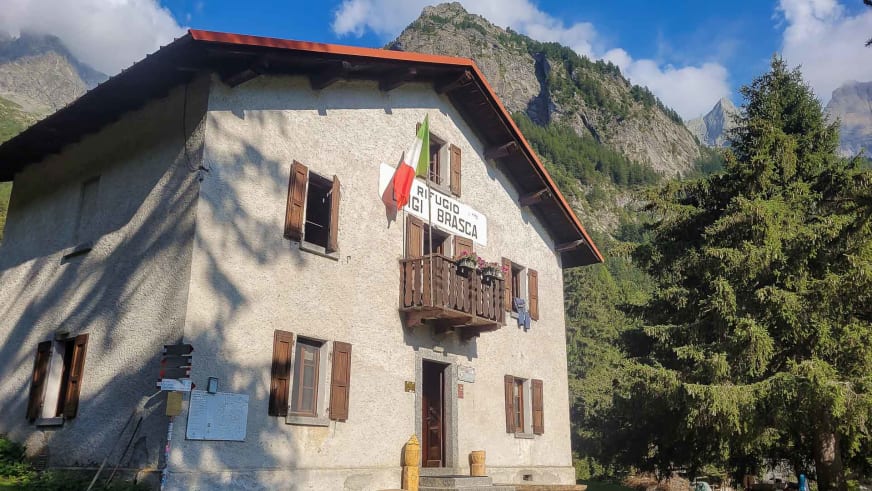 Außenansicht des Rifugio Brasca, einer zweistöckigen Berghütte mit heller Fassade und roten Ziegeldach in alpiner Umgebu