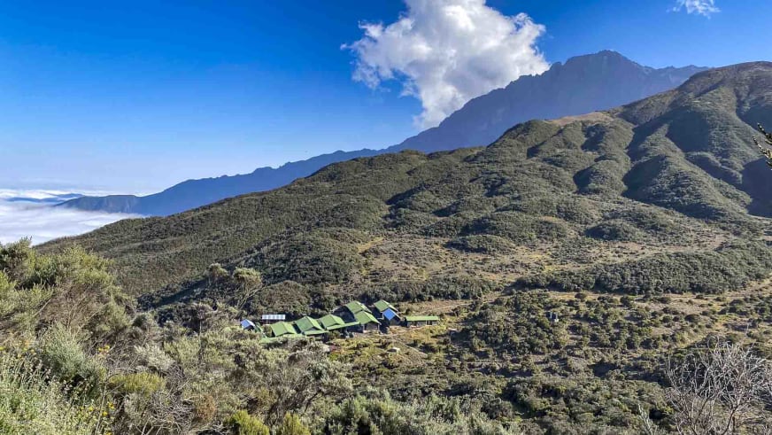 Panoramablick auf die Sattelhütte (Mt. Meru) mit grünen Zelten und Gebäuden zwischen niedriger Bergvegetation.