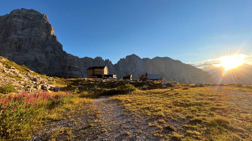 Das Rifugio Brentei bei Sonnenuntergang mit goldenen Felswänden und blühenden Alpenblumen im Vordergrund.