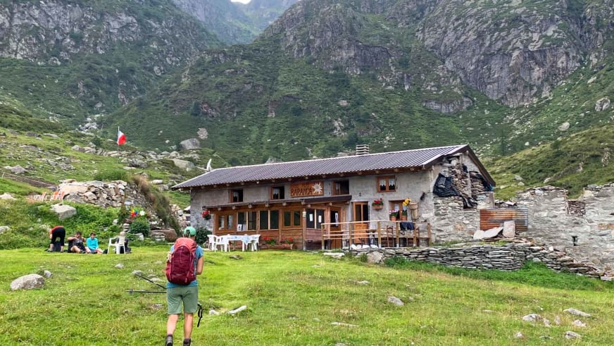 Wanderer mit rotem Rucksack vor dem Rifugio Alpe Baranca auf grüner Almwiese in den Walliser Alpen.