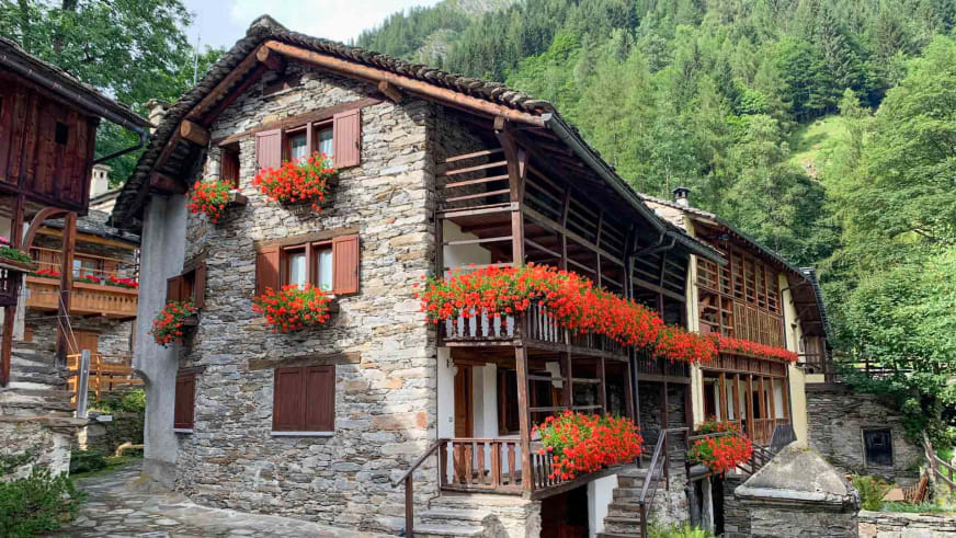 Traditionelle Steinhäuser mit Holzbalkonen und roten Geranien umgeben das Rifugio Brusà in alpiner Landschaft.