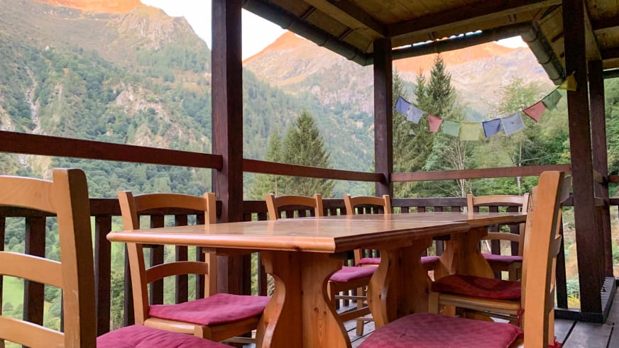 Überdachte Terrasse des Rifugio Brusà mit ovalem Holztisch und Blick auf bewaldete Berghänge.