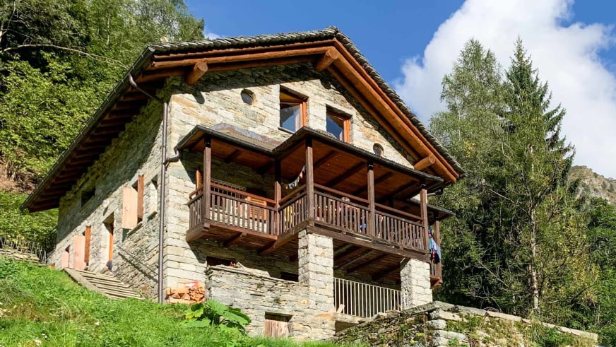 Das Rifugio Brusà zeigt sich als traditionelles Steinhaus mit Holzbalkon auf grüner Hangwiese im Wald.