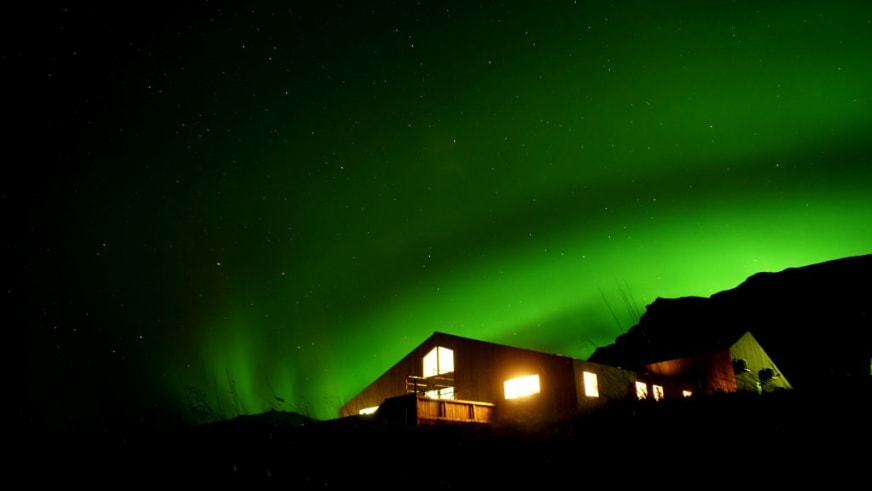 Die Holy Ram Lodge bei Nacht unter grünen Polarlichtern vor der Bergkulisse von Snæfellsnes mit warm erleuchteten Fenste