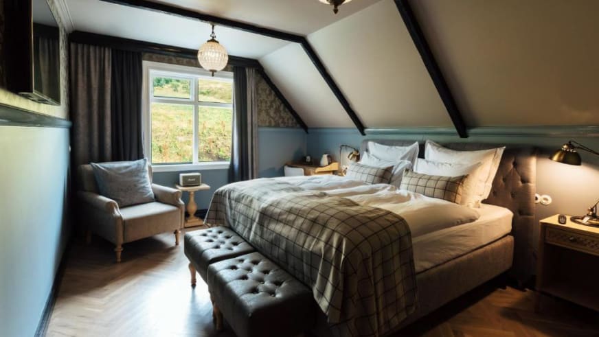 Gemütliches Doppelzimmer in der Holy Ram Lodge mit sichtbaren Holzbalken, karierter Bettwäsche und Polstersessel am Fens