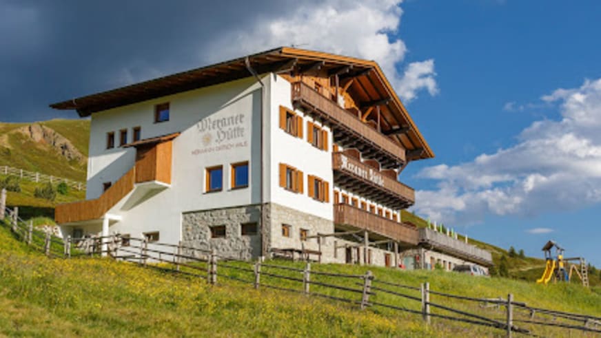 Die Meraner Hütte zeigt sich mit weißer Fassade, Holzbalkonen und traditionellem Satteldach auf grüner Almwiese.