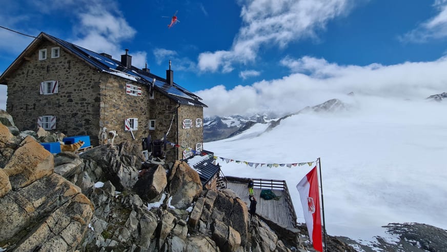 Das Brandenburger Haus auf einem Felsgrat mit rotem Hubschrauber am Himmel über dem Gletscherplateau.