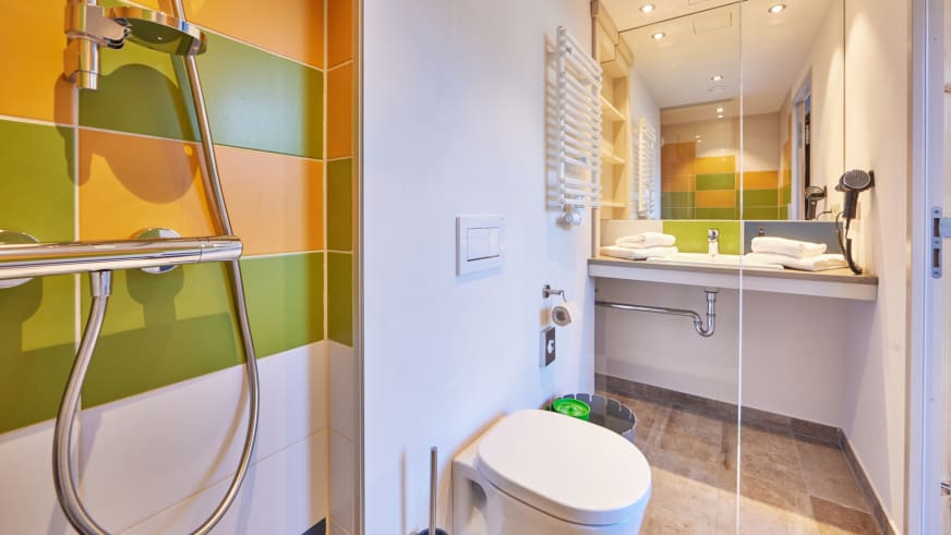 Badezimmer im Explorer Hotel Stubaital mit farbiger Wandgestaltung und Glasdusche.
