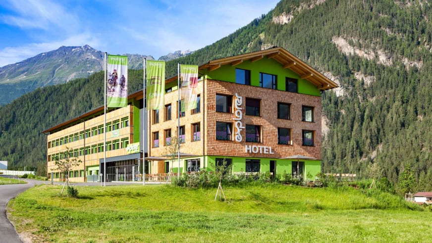 Außenansicht Explorer Hotel Stubaital im Sommer mit bewaldeten Bergen im Hintergrund.