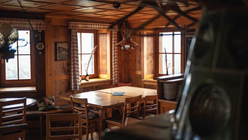 Gemütlicher Gastraum der Leutkircher Hütte mit Holztisch, karierten Vorhängen und Blick auf verschneite Berglandschaft.