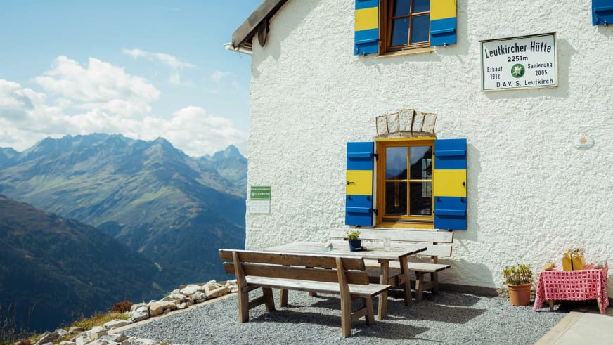 Die Leutkircher Hütte zeigt sich mit weißer Fassade und blauen Fensterläden vor weitem Bergpanorama der Lechtaler Alpen.