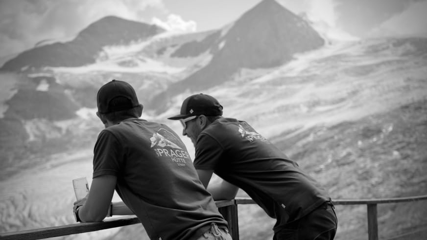 Zwei Bergsteiger auf der Terrasse der Neuen Prager Hütte blicken über das verschneite Bergpanorama der Hohen Tauern.