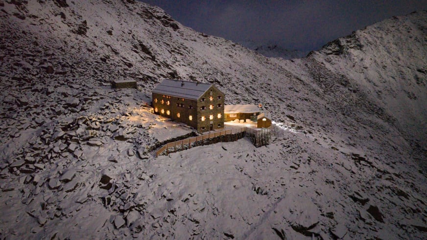 Die Neue Prager Hütte steht nachts mit warmer Fensterbeleuchtung in der verschneiten Hochgebirgslandschaft auf 2.796 m.