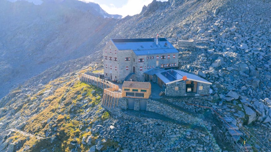 Luftaufnahme der Neuen Prager Hütte mit Hauptgebäude und Anbauten auf einem Geröllplateau in hochalpinem Gelände.