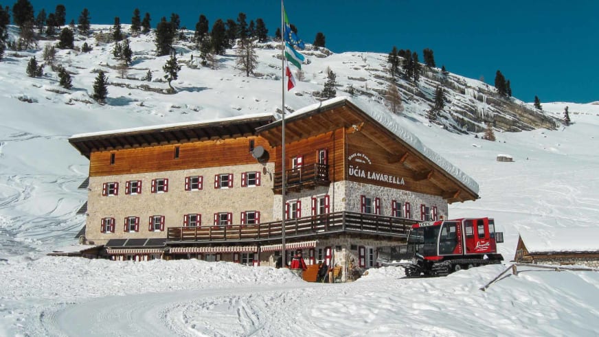 Das Rifugio Lavarella zeigt sich im Winter mit tiefem Neuschnee, roter Pistenraupe und typischer Alpenarchitektur.