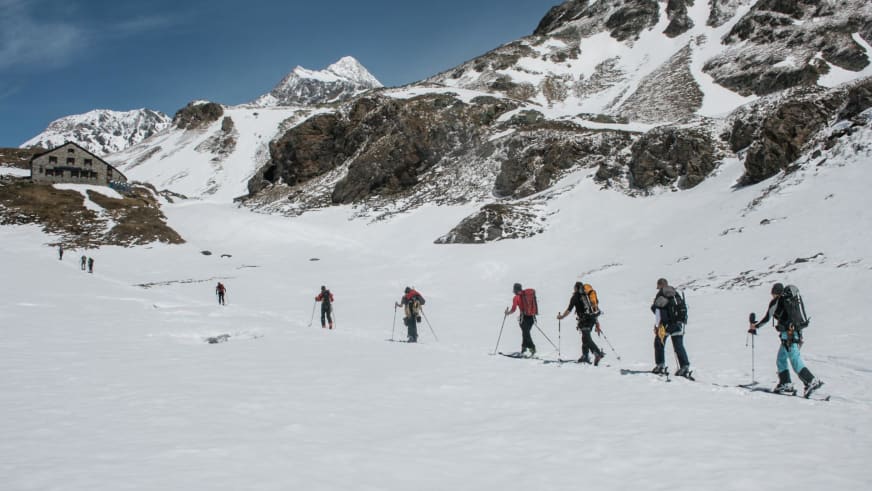 Zehn Skitourengeher steigen durch Tiefschnee zur Cabane de Chanrion auf, die Berghütte ist links im Bild sichtbar.