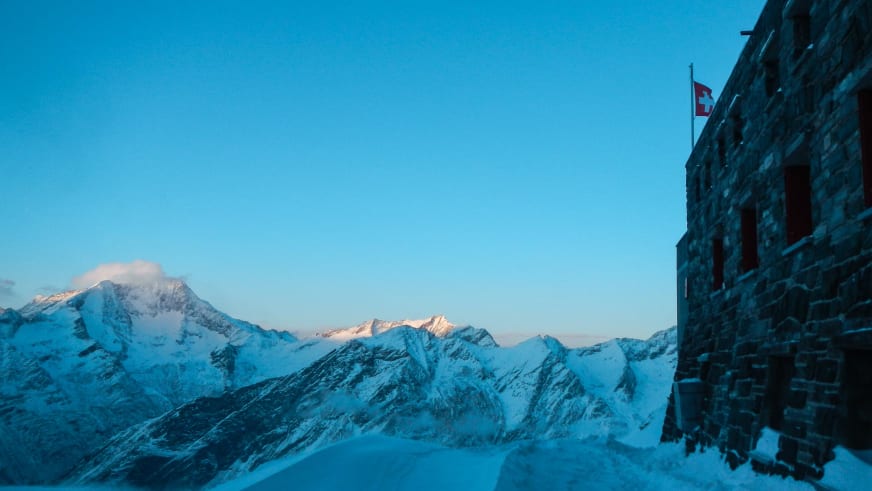 Die Britannia-Hütte aus Naturstein steht verschneit auf einem Felsplateau mit Schweizer Flagge in der Hochgebirgslandschaft.
