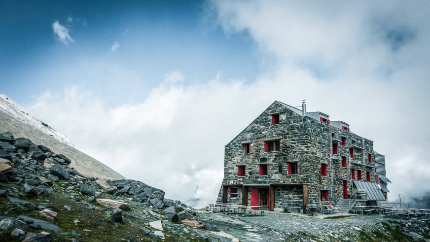 Außenansicht der Britannia-Hütte aus grauem Naturstein mit roten Fensterläden auf felsigem Untergrund im Sommer.