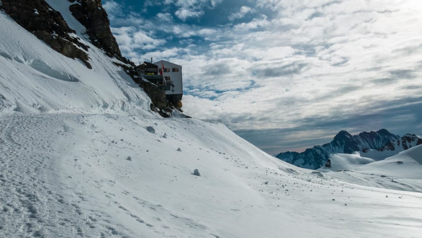 Die Mönchsjochhütte steht auf einem Felssockel umgeben von Schneehängen und schneebedeckten Viertausendern der Berner Alpen.