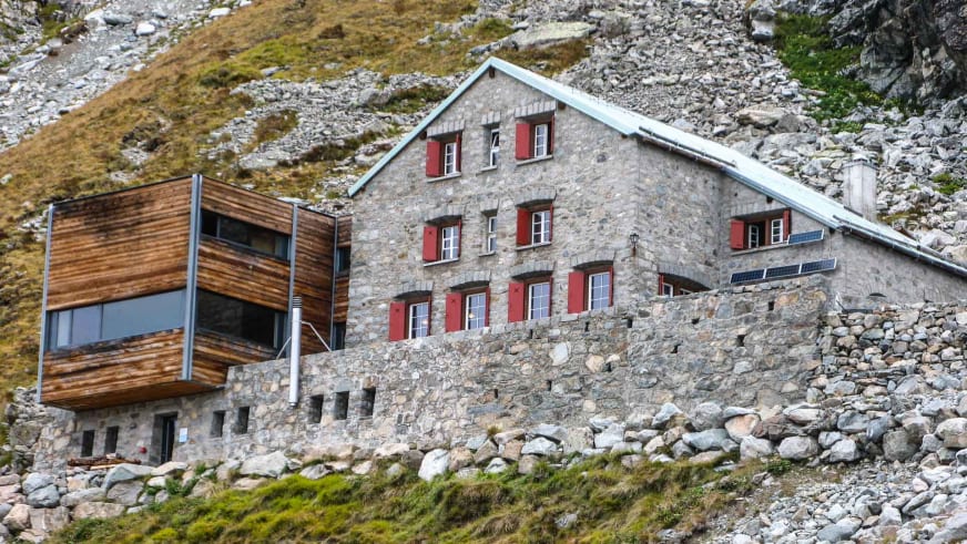 Außenansicht der Tschiervahütte mit Steinbau, roten Fensterläden und Holzanbau auf Geröllhang in 2583 m Höhe.