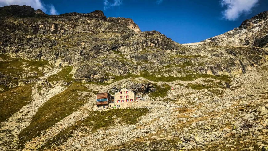 Panoramaaufnahme der Tschiervahütte auf einem Plateau umgeben von Geröllfeldern mit Felswand im Hintergrund der Bernina-Alpen.