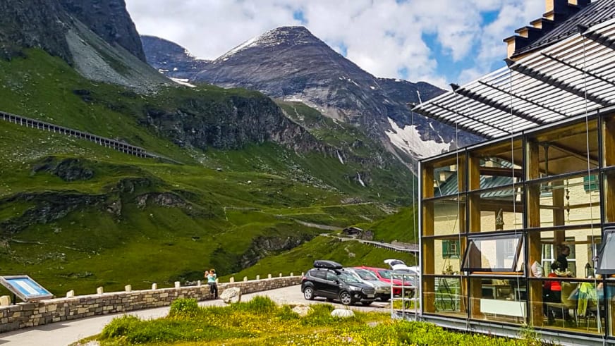 Der moderne Glasanbau des Glocknerhaus steht vor der Bergkulisse der Hohe Tauern mit Parkplatz und Steinmauer.