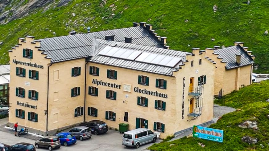 Luftaufnahme zeigt das Glocknerhaus mit gelber Fassade, Restaurant und Parkplatz umgeben von grünen Almwiesen in Österreich.
