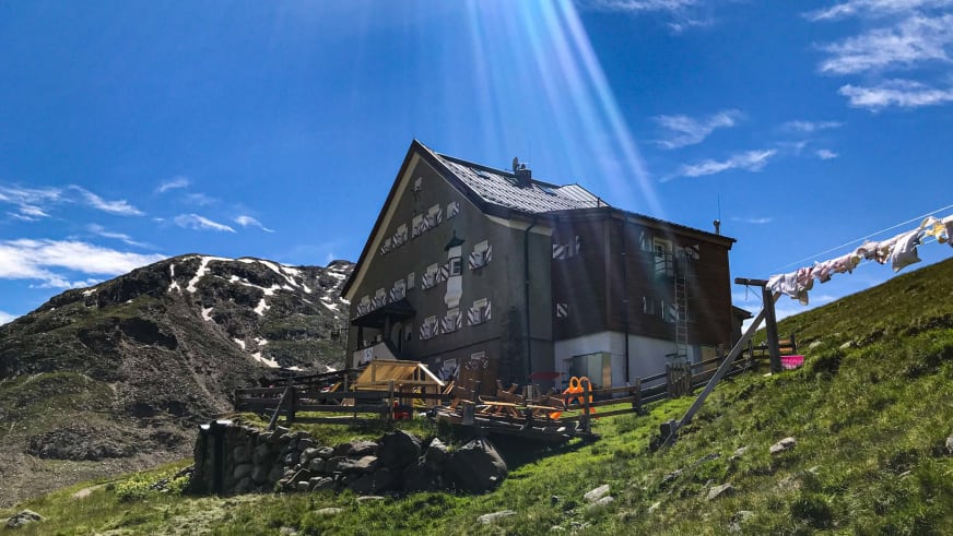 Das Hochjoch Hospiz zeigt sich als alpine Berghütte mit Holzterrasse auf grüner Almwiese zwischen Felsblöcken.