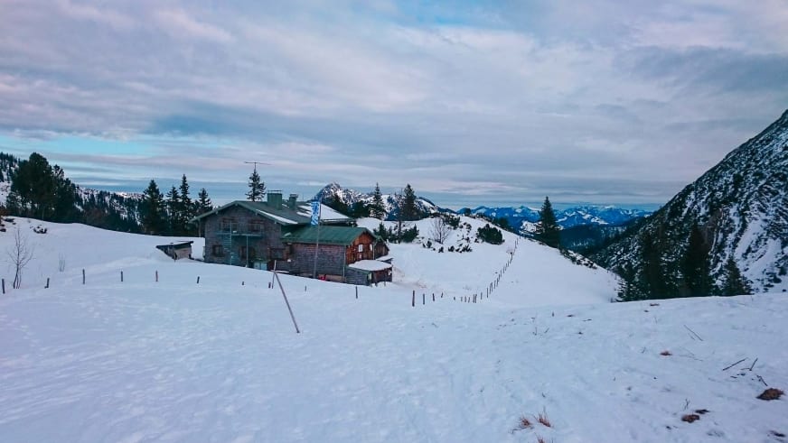 Das Taubensteinhaus steht im Winter auf einem verschneiten Plateau in den Bayerischen Voralpen mit Bergpanorama.