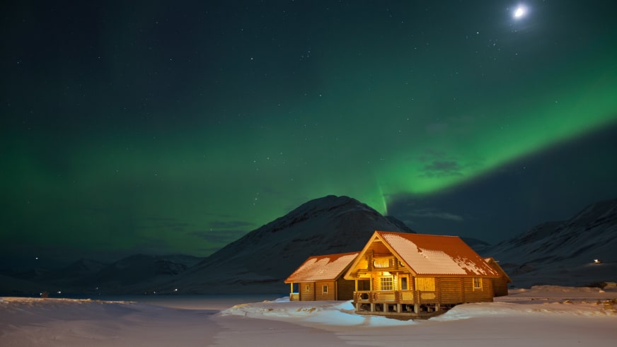 Das beleuchtete Brimnes Hotel and Cabins steht unter grünen Nordlichtern in verschneiter Landschaft Islands.