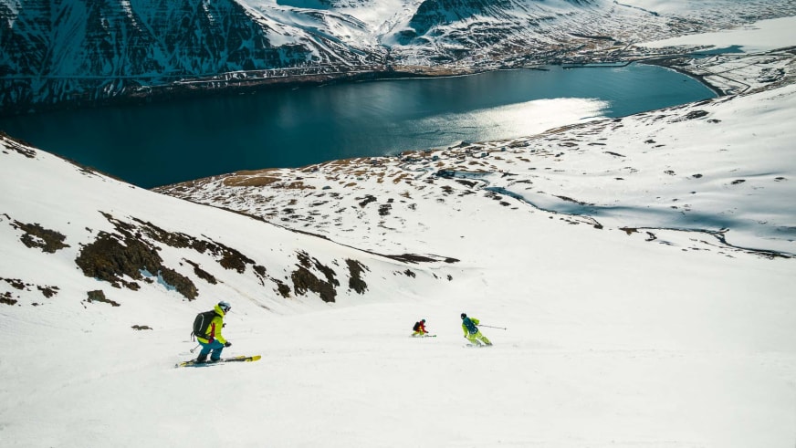 Drei Skifahrer bei Firnabfahrt zum Fjord nahe Brimnes Hotel and Cabins in Islands Winterlandschaft.