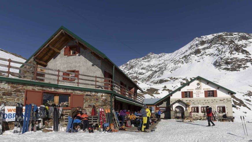 Zahlreiche Skitourengeher pausieren auf der Holzterrasse des Rifugio Branca mit Skiausrüstung bei Sonnenschein.