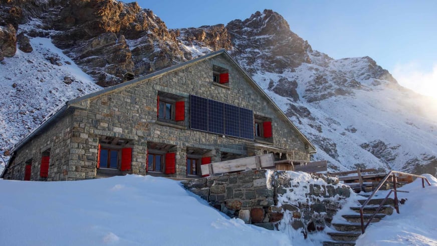 Außenansicht der Schönbielhütte im Winter mit Natursteinfassade und roten Fensterläden vor verschneiten Gipfeln.