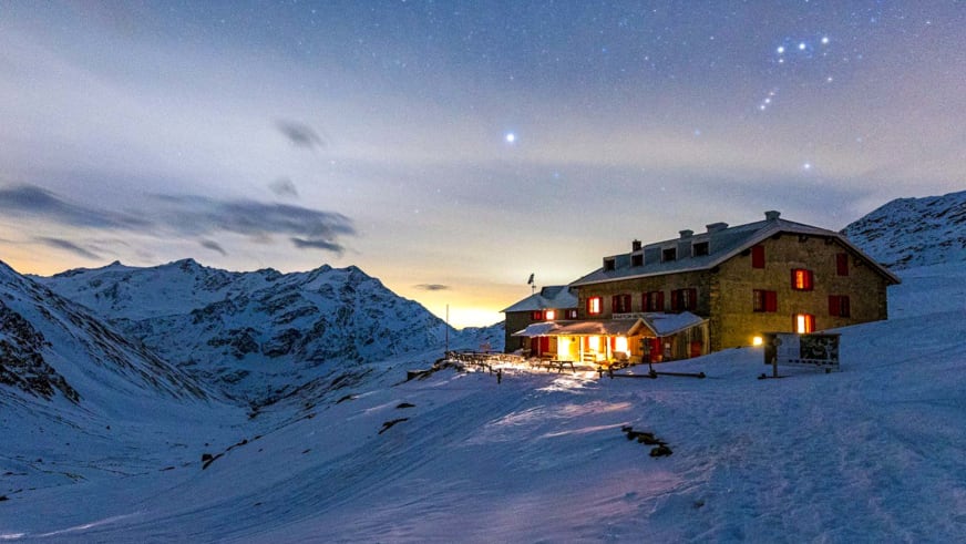 Das Rifugio Pizzini zeigt sich bei Nacht mit warmer Innenbeleuchtung in tiefverschneiter Hochgebirgslandschaft auf 2706 m.
