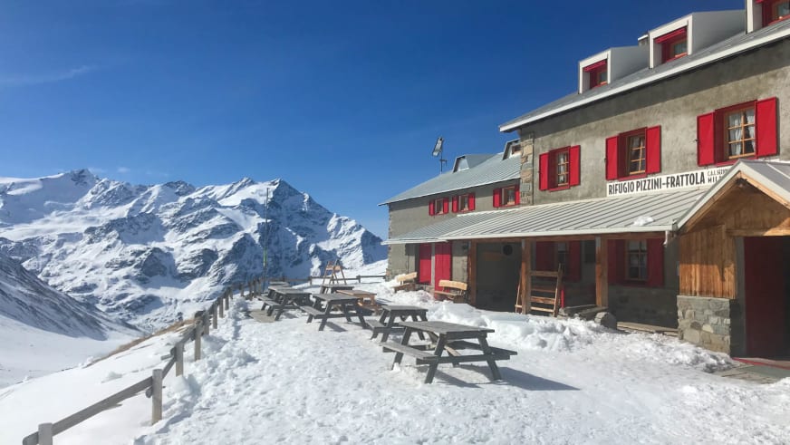 Blick auf das Rifugio Pizzini mit Holzterrasse und Sitzbänken vor verschneiter Bergkulisse bei klarem Winterwetter.