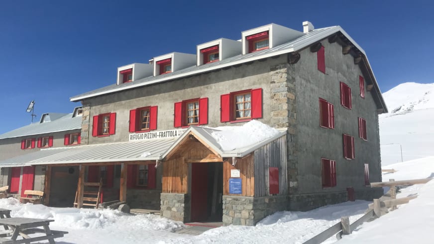 Außenansicht des Rifugio Pizzini mit traditioneller Steinarchitektur, roten Fensterläden und Holzterrasse im Schnee.
