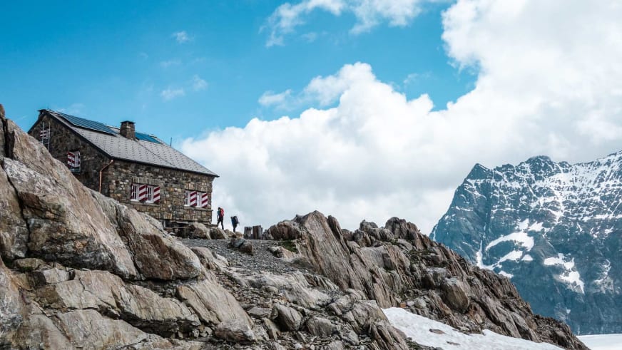 Die Tierberglihütte steht auf felsigem Terrain mit zwei Wanderern vor schneebedeckten Gipfeln der Urner Alpen.